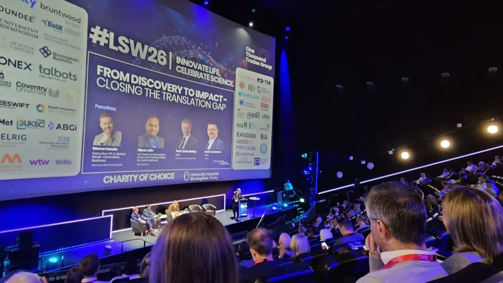 Speakers on stage for the From Discovery to Impact panel at Life Sciences Week 2026, with a full audience in front of the big screen