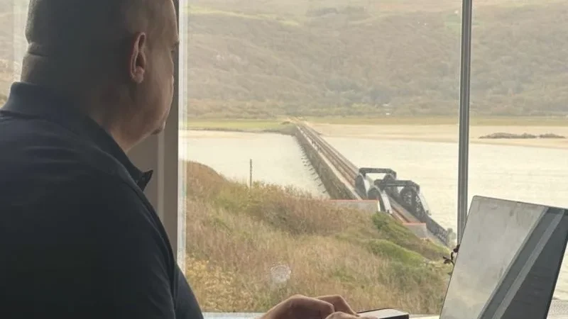 Paul Cadman working on a laptop by a window overlooking Barmouth Bridge