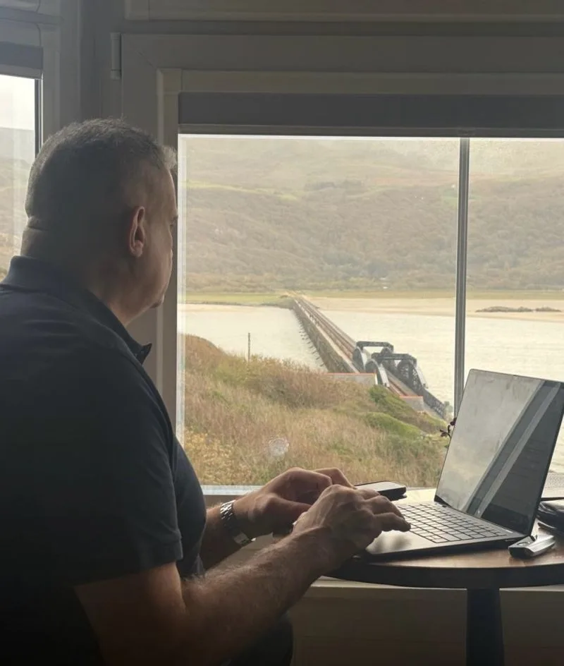 Paul Cadman working on a laptop by a window overlooking Barmouth Bridge